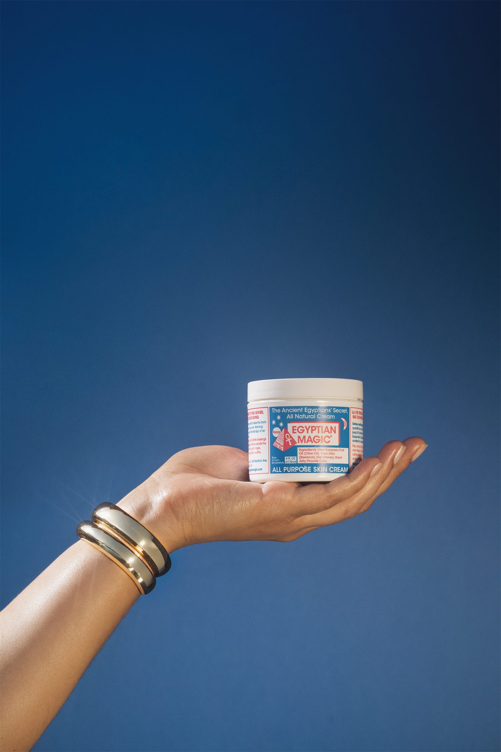 A jar of Egyptian Magic offered on an open palm with gold bangles, against a deep cobalt sky