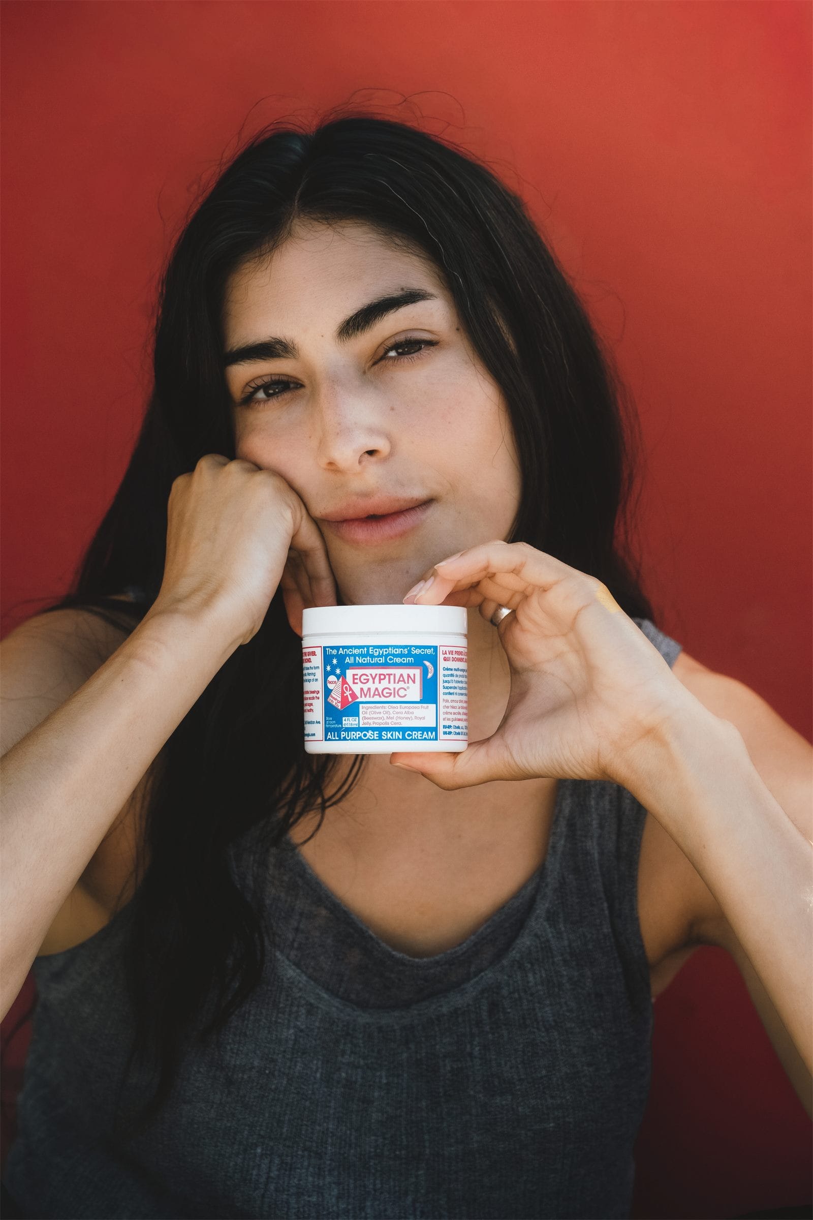 Model presenting a jar of Egyptian Magic to camera against a deep red wall